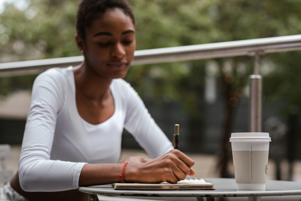 African American woman writing in a notebook at a cafe table outdoors with a coffee.