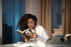 Home Young woman with curly hair engrossed in a book beside a typewriter at home.