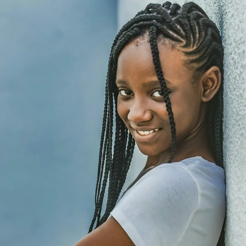 A cheerful image of a teenage girl with braided hair, smiling against a wall outdoors.
